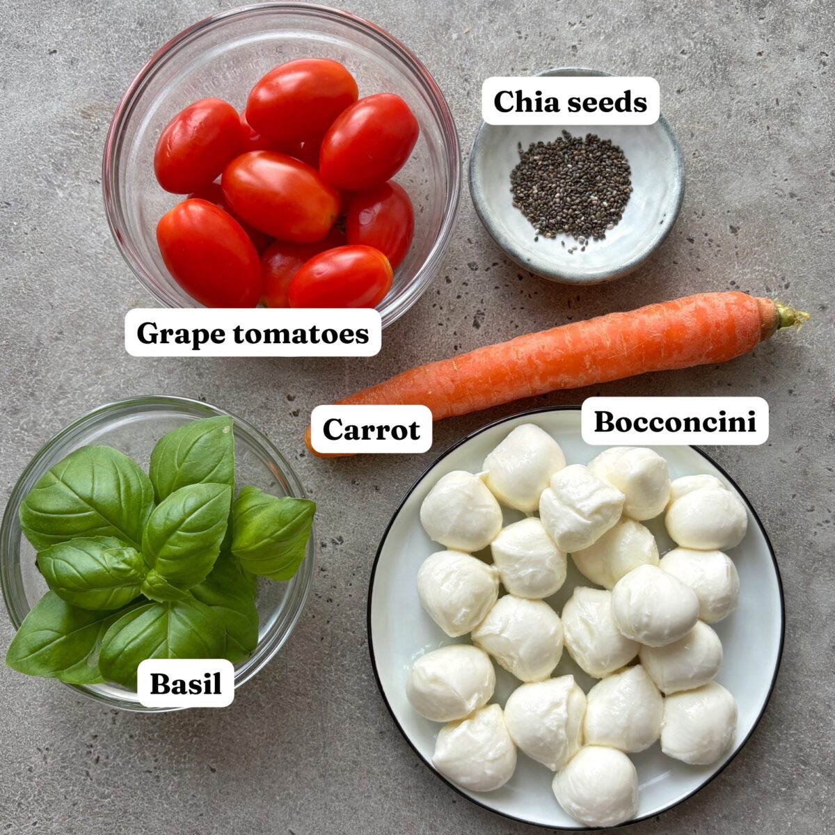 Ingredients on a grey surface including grape tomatoes, chia seeds, a carrot, basil leaves, and a bocconcini cheese.