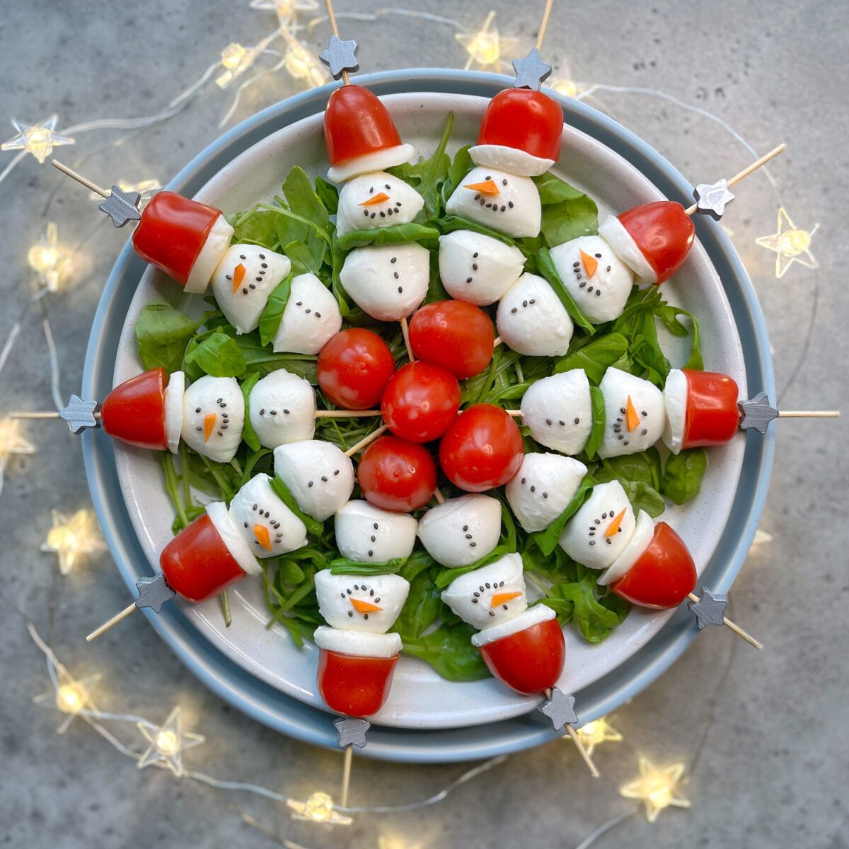 A plate of salad with skewers arranged in a circle, each decorated to look like snowmen using mozzarella, tomatoes, and arugula. A festive string of lights surrounds the plate.