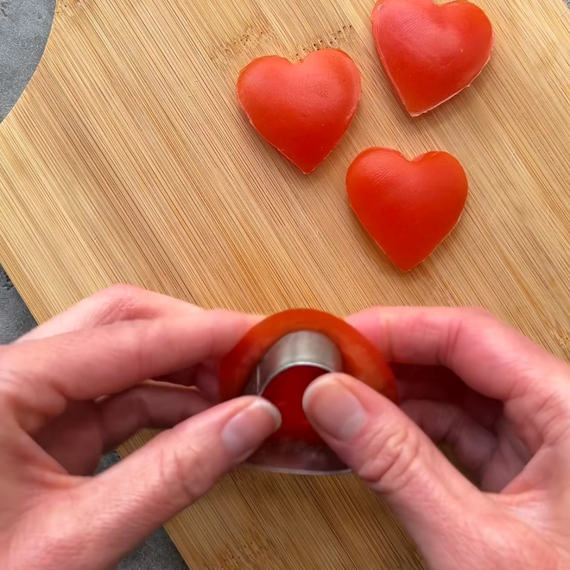 Hands using a heart-shaped cutter to make heart-shaped pieces of tomato.