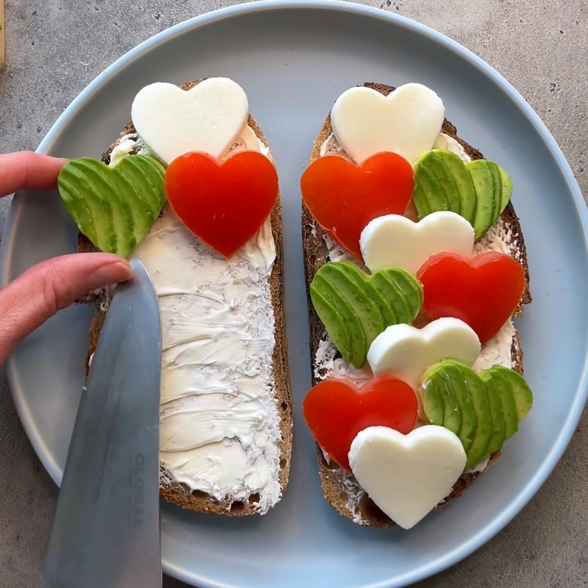 Avocado Cheese Toast: Two slices of bread with cream cheese, topped with heart-shaped mozzarella, tomato, and avocado on a plate. A hand is spreading creamy cheese on one slice.