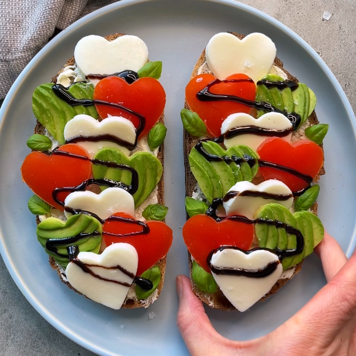 Avocado Cheese Toast featuring two slices of bread topped with heart-shaped mozzarella, tomato slices, avocado, basil leaves, and a drizzle of balsamic glaze on a gray plate.