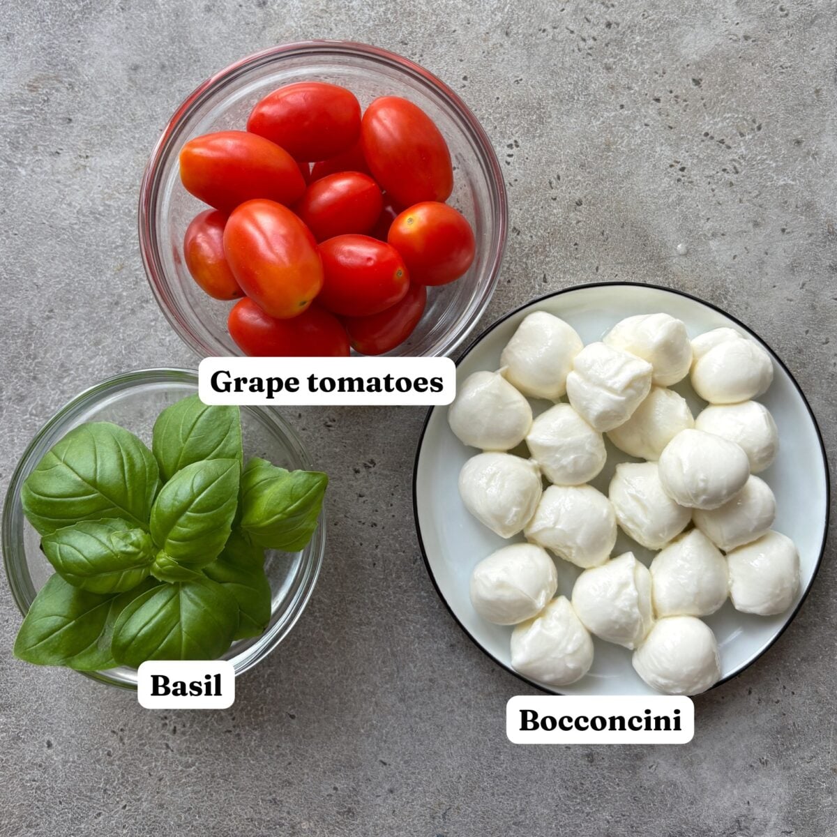 Three bowls on a gray surface containing grape tomatoes, fresh basil leaves, and bocconcini mozzarella balls.