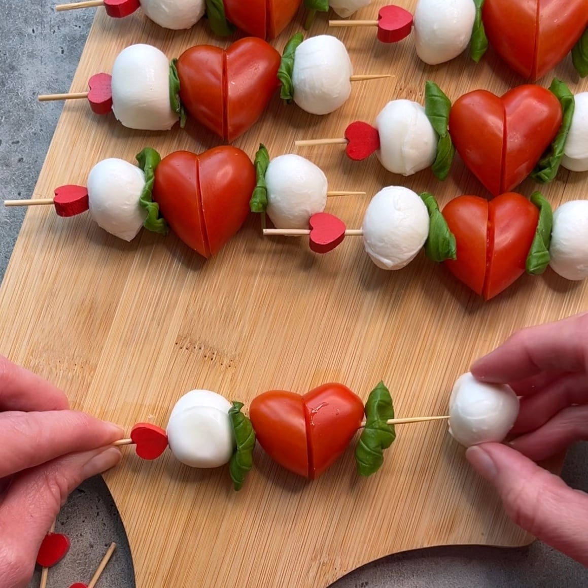 Caprese Skewers with mozzarella balls, basil leaves, and tomato halves shaped like hearts are arranged on a wooden board.