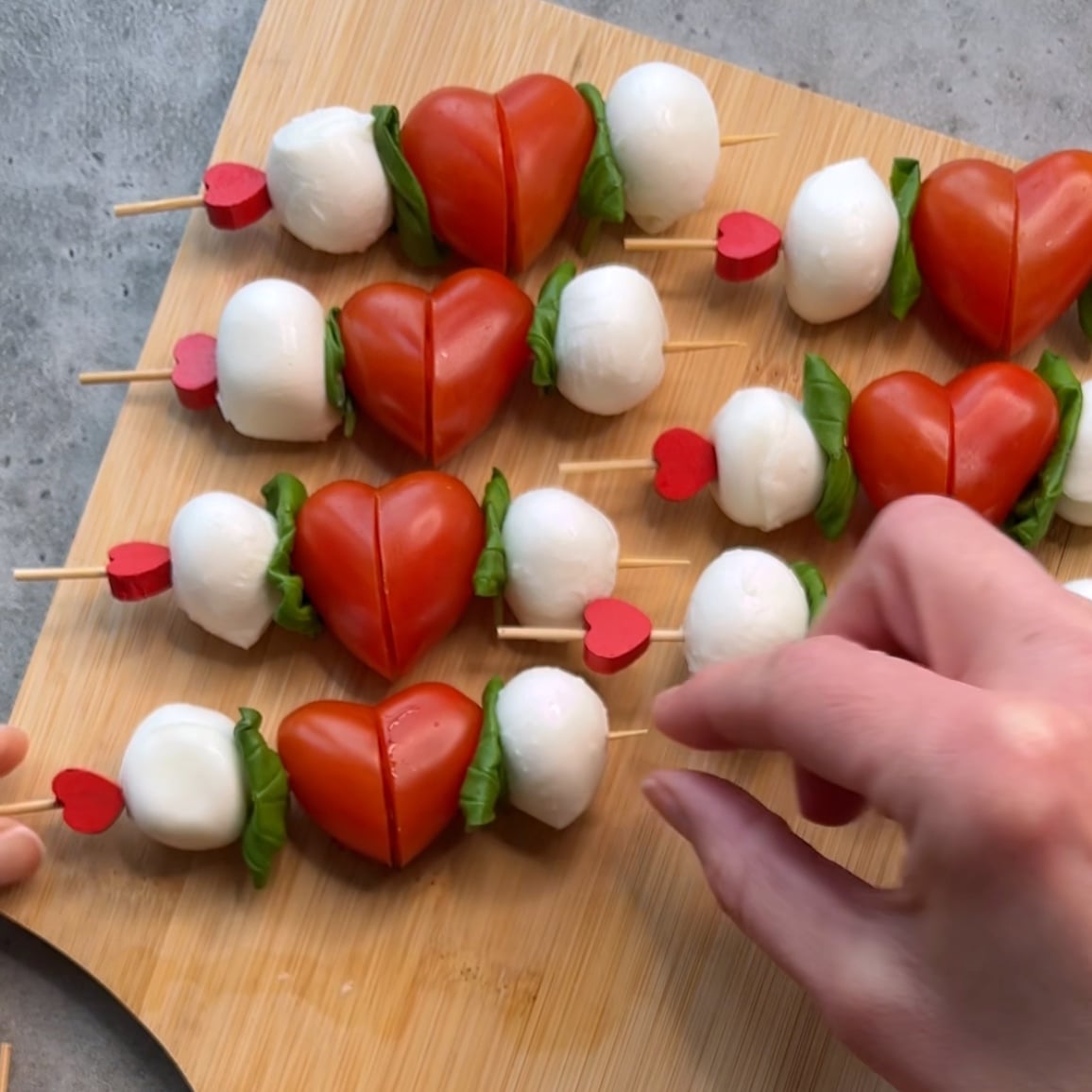 Caprese Skewers with mozzarella balls, basil leaves, and halved tomatoes shaped like hearts are arranged on a wooden board, with a hand reaching towards them.