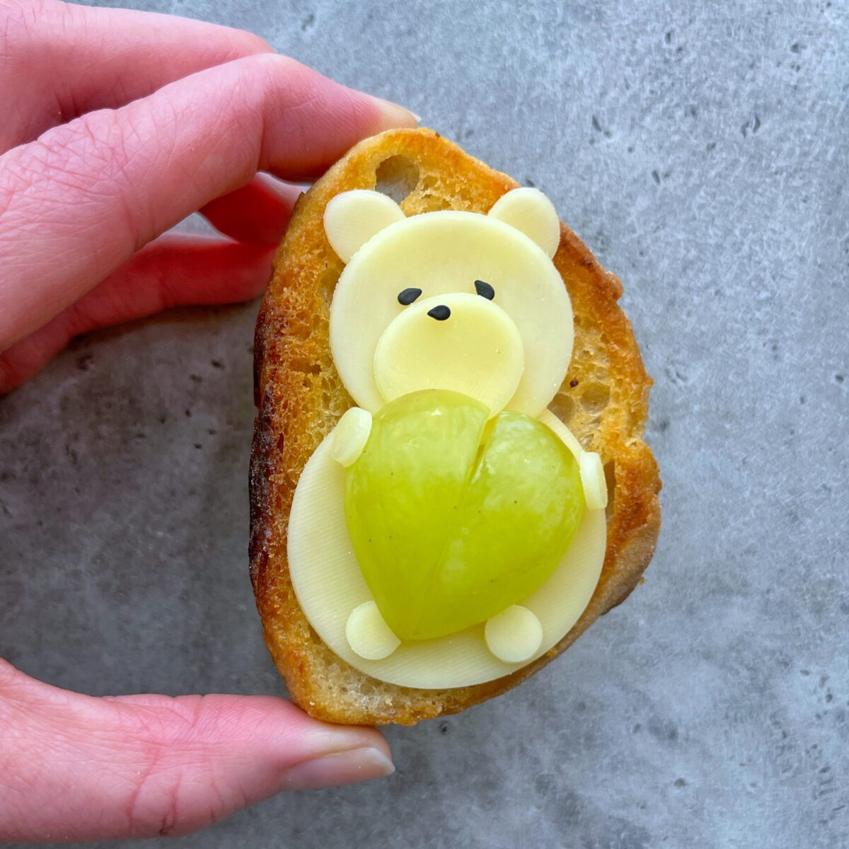 A hand holds a slice of bread topped with cheese shaped like a bear and a green grape arranged as a heart.
