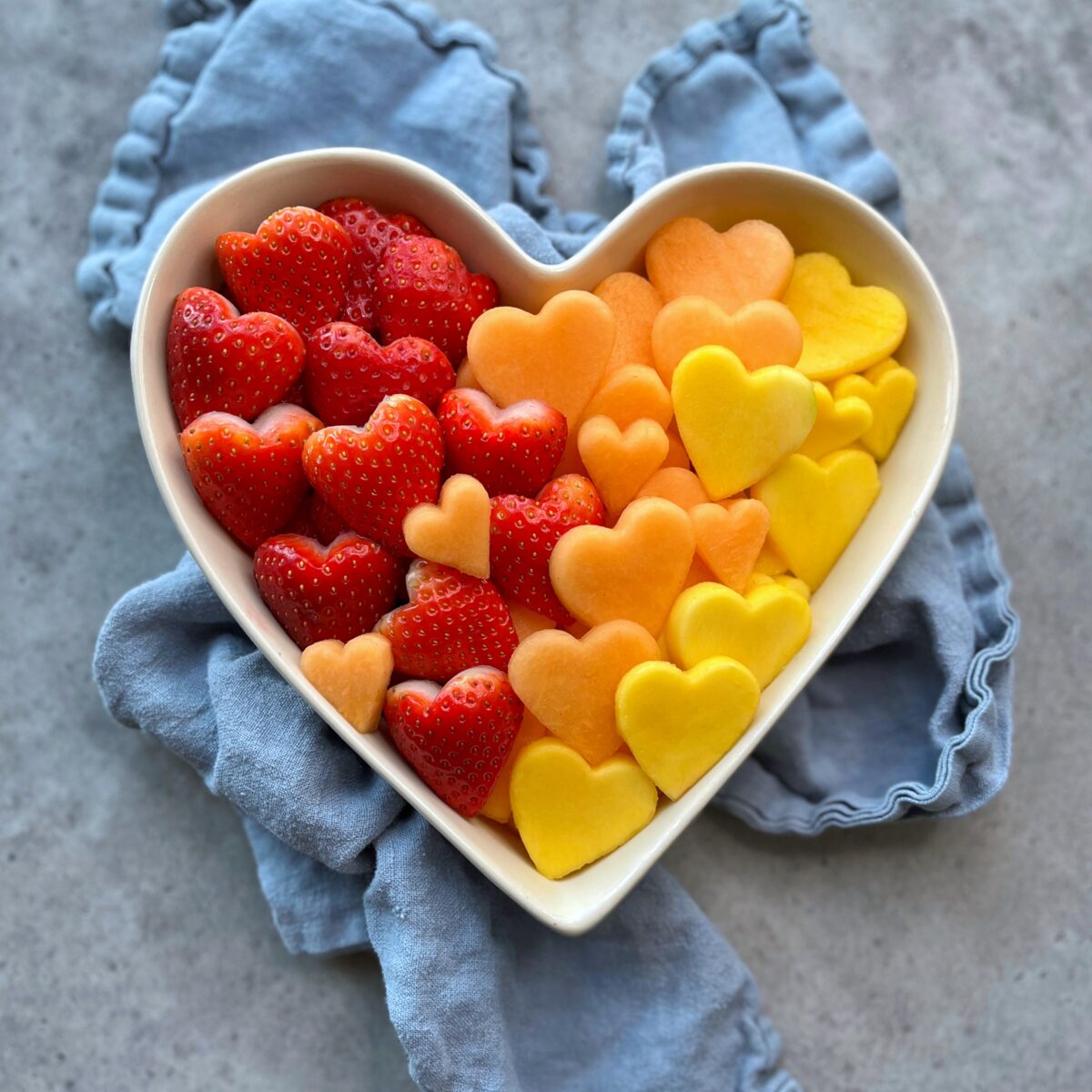A heart-shaped bowl filled with heart-shaped bites of strawberries, cantaloupe, and mango, arranged on a blue cloth.