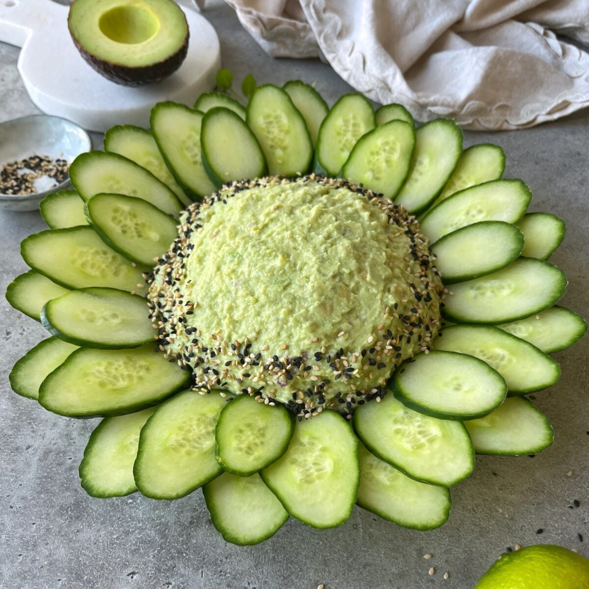 A dome of guacamole dip surrounded by cucumber slices arranged in a circular pattern, garnished with mixed seeds, on a gray surface.