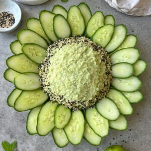 A round platter of guacamole dip is surrounded by cucumber slices arranged in a flower pattern, with seasoning sprinkled around the dip&rsquo;s edge, all displayed on a gray surface.
