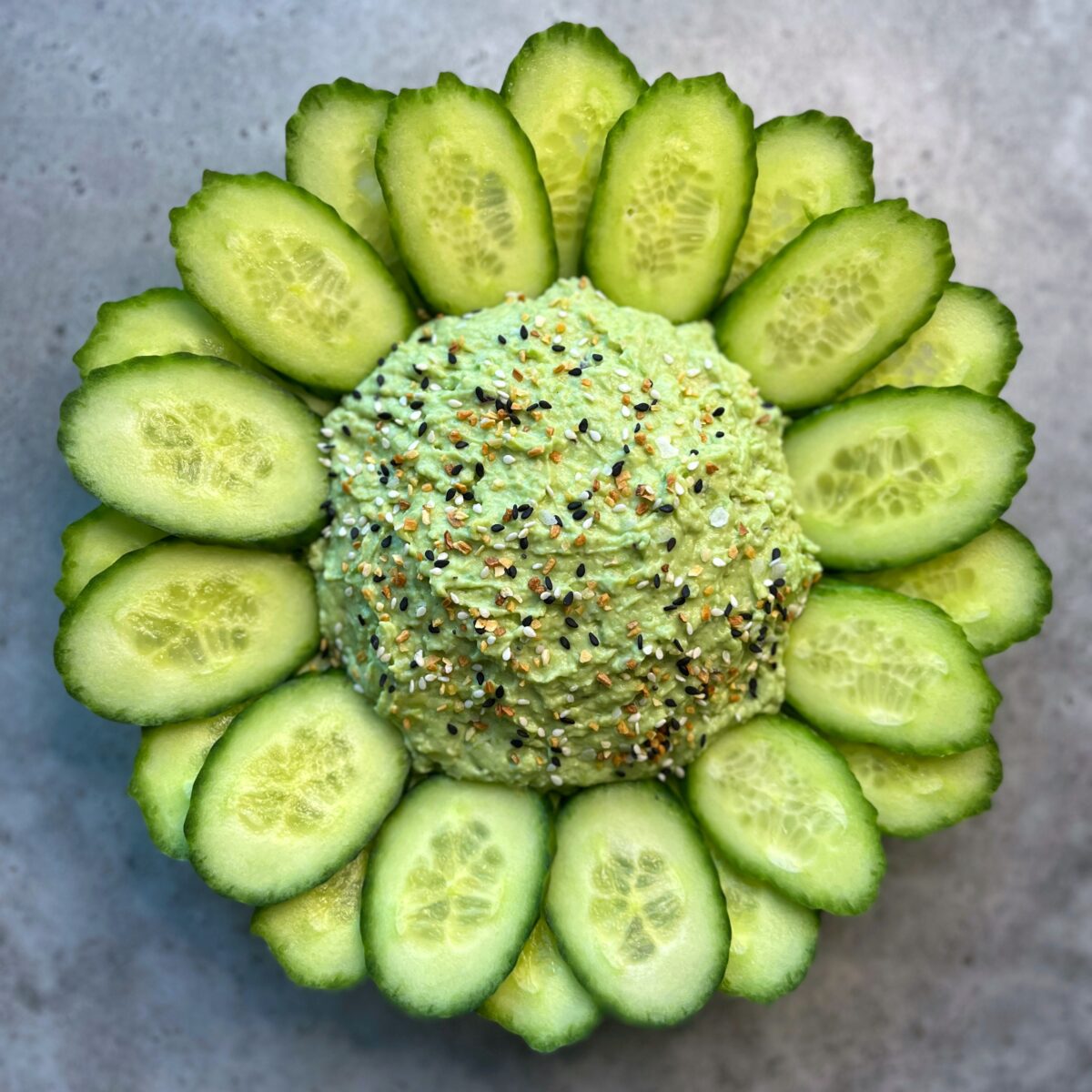 Full view of a mound of avocado dip garnished with seasoning, surrounded by neatly arranged cucumber slices on a gray surface.