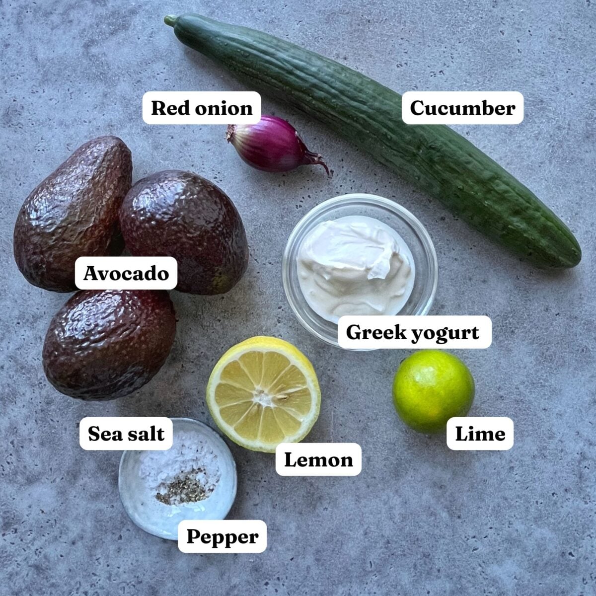 Ingredients for Guacamole Dip arranged on a gray surface: red onion, cucumber, avocado, Greek yogurt, sea salt, pepper, lemon, and lime, each labeled.