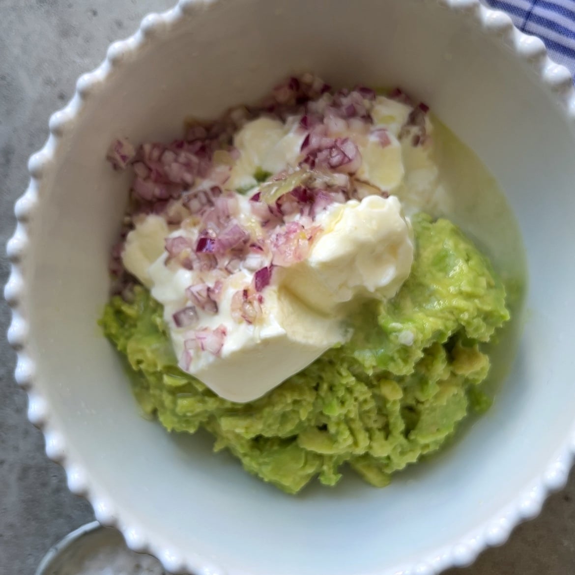 A white bowl containing Guacamole Dip made with mashed avocado, Greek yogurt, and finely chopped red onions on a light surface.