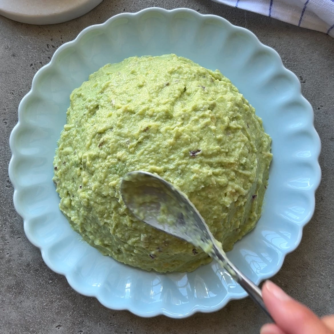 A hand holds a spoon, spreading a mound of green Guacamole Dip mixture on a scalloped plate.