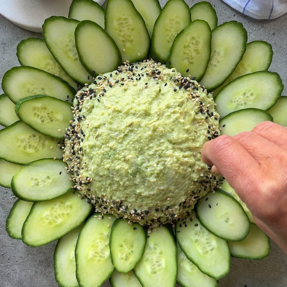 A hand sprinkles seasoning on a mound of guacamole dip, surrounded by neatly arranged cucumber slices on a round plate.