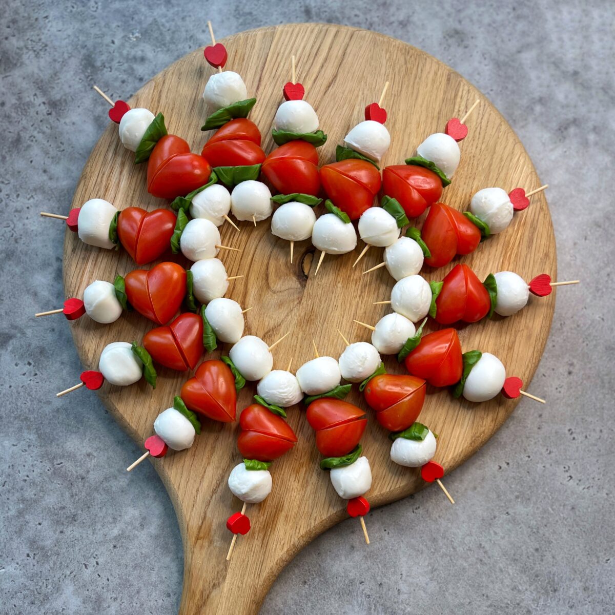 A wooden board displays delicious bites with mozzarella balls, cherry tomatoes, and basil leaves arranged in a circular pattern on a gray surface.