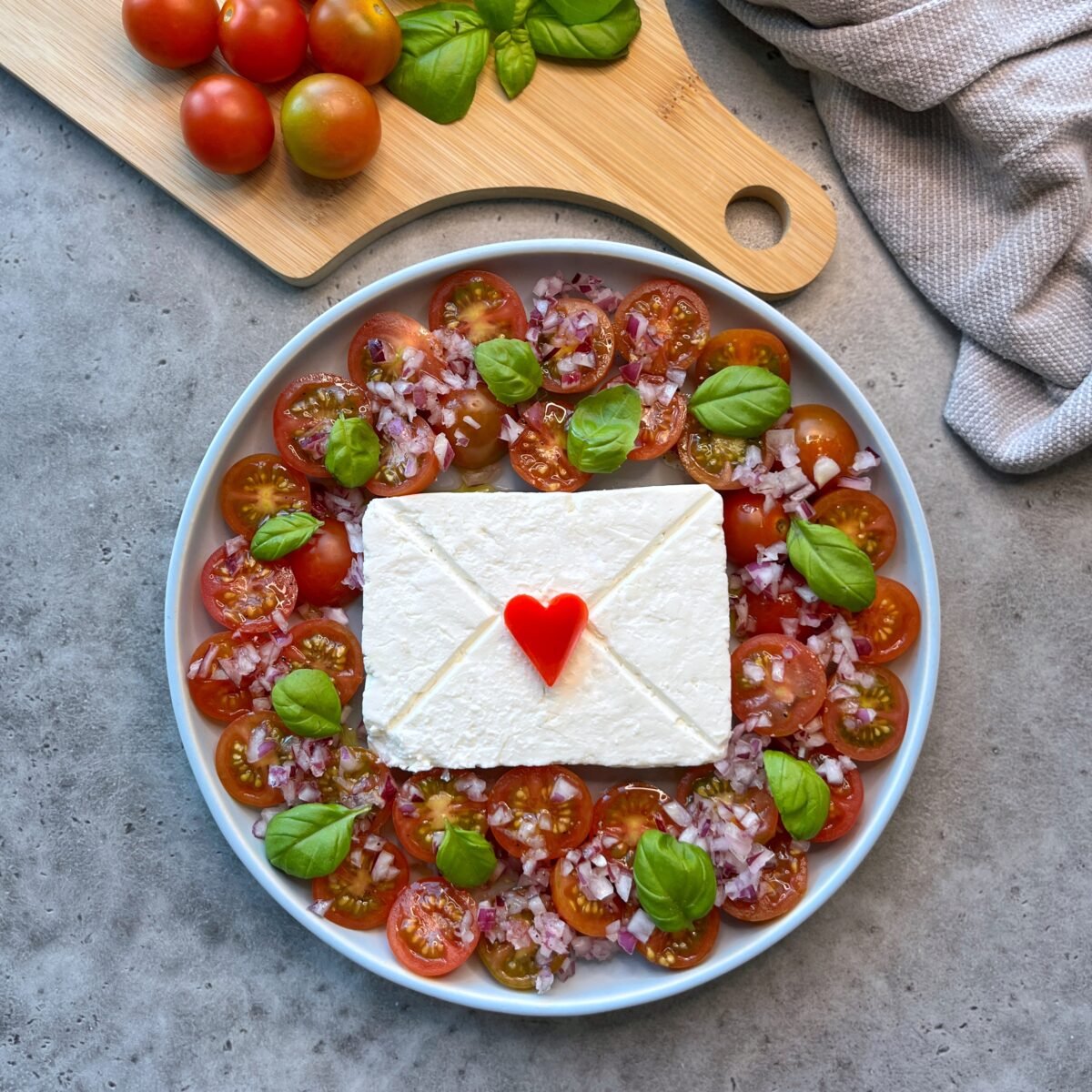 A plate with sliced cherry tomatoes, feta cheese shaped like an envelope with a red heart, chopped onions, and basil leaves sits next to a cutting board with tomatoes and basil.
