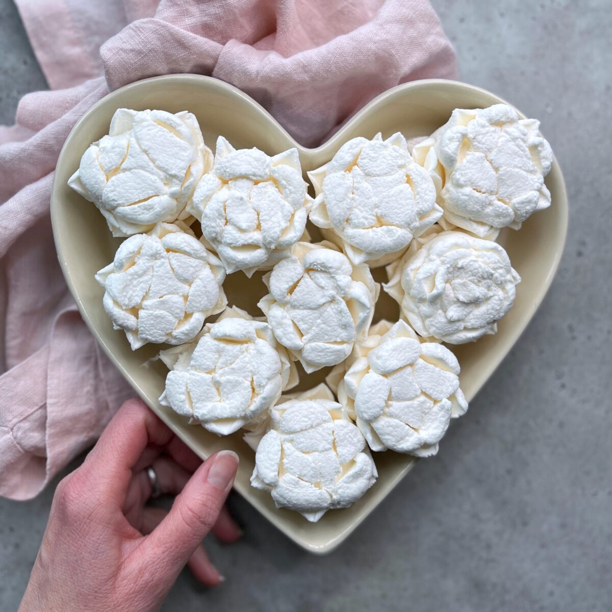 A hand holds a heart-shaped bowl filled with white, flower-shaped meringue bites. A light pink cloth is partially visible beneath the bowl, set on a gray surface—a charming touch for those who love sweet food recipes.