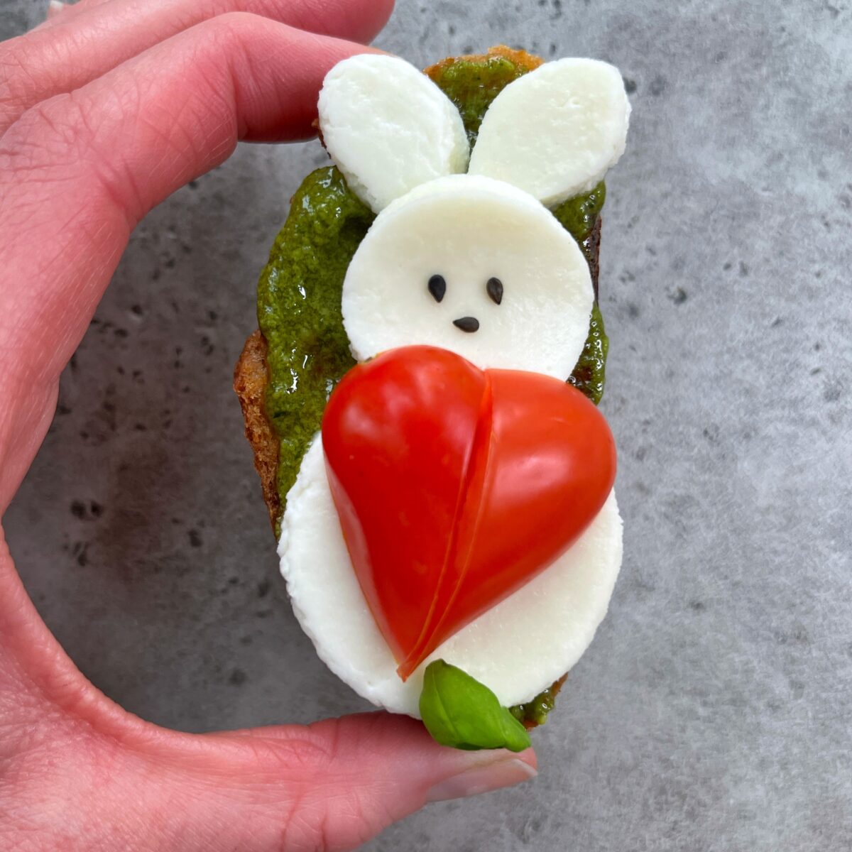 A hand holds a piece of bread topped with pesto, mozzarella slices shaped like a bunny, black sesame seeds for eyes, and a halved tomato forming a heart on the bunny’s chest—perfect for creative food recipes and healthy bites.