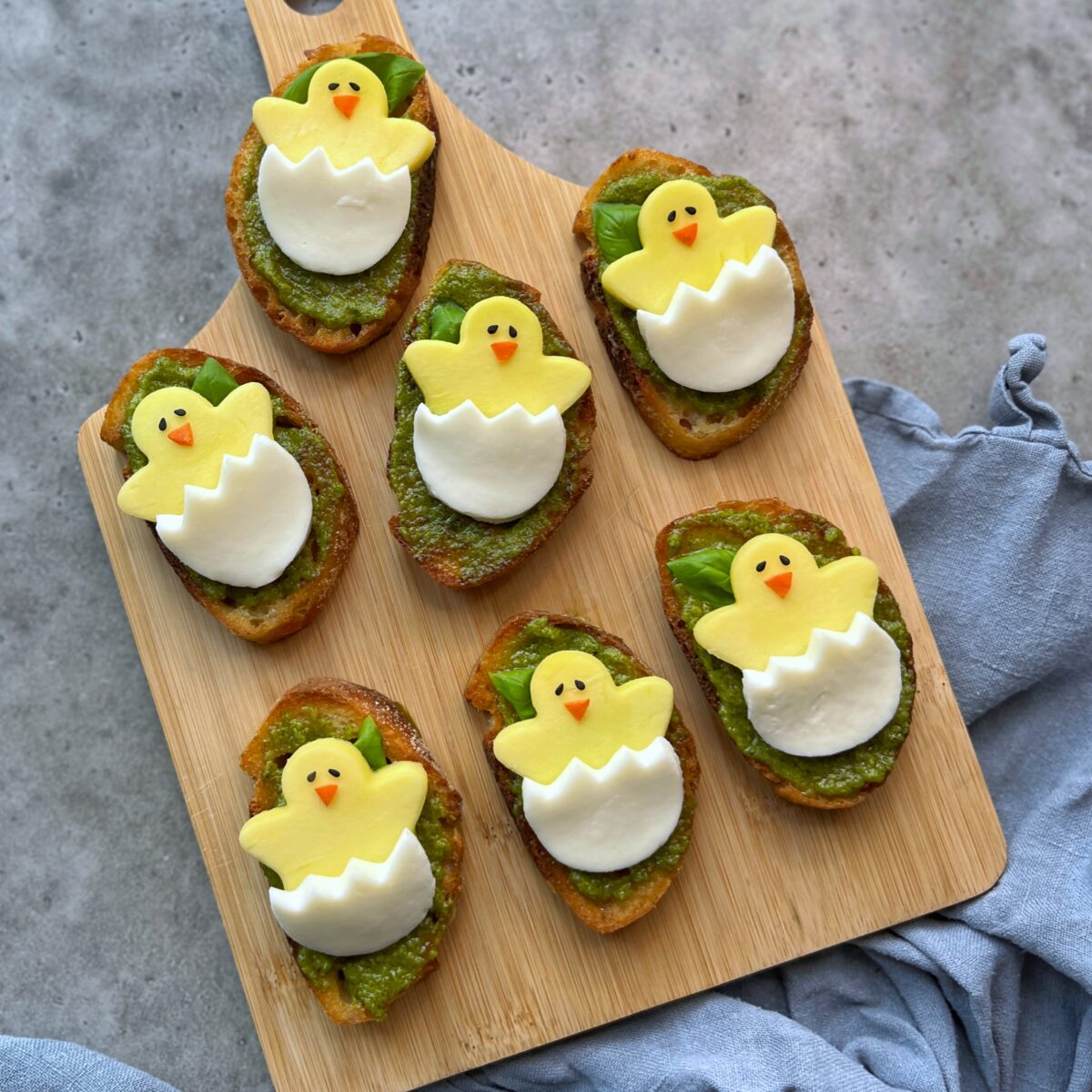 Seven pieces of crostini topped with pesto and cheese and mango slices shaped like hatching chicks.