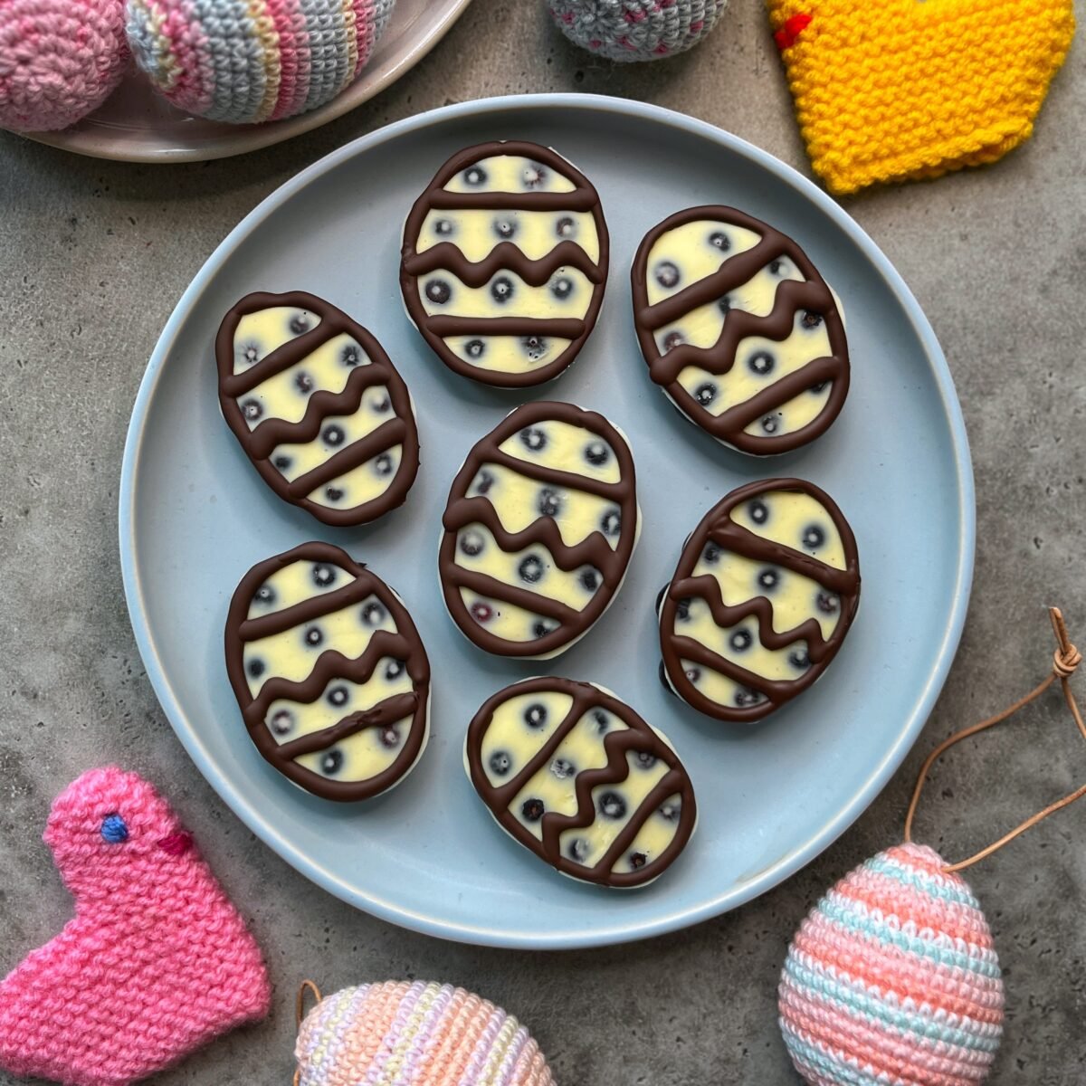 A plate of seven chocolate Easter eggs with white and dark chocolate decoration, surrounded by knitted and crocheted Easter decorations.
