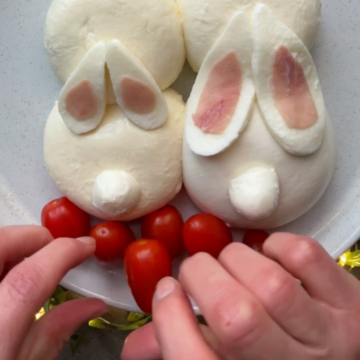 A hand places grape tomatoes on a platter with burrata bunnies in the center.