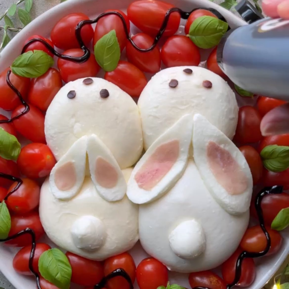 A festive Easter Salad features burrata balls shaped like rabbits, nestled among cherry tomatoes, basil leaves, and finished with a drizzle of balsamic glaze and olive oil.