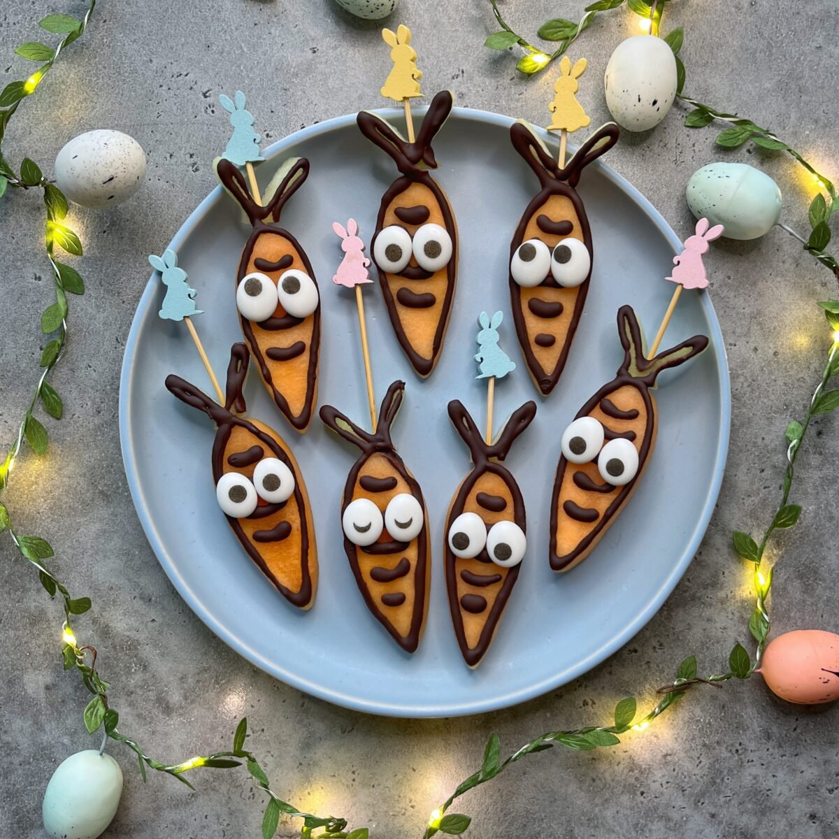 A plate of seven carrot-shaped fruit snacks with googly eyes and chocolate details on sticks, surrounded by pastel eggs and string lights.