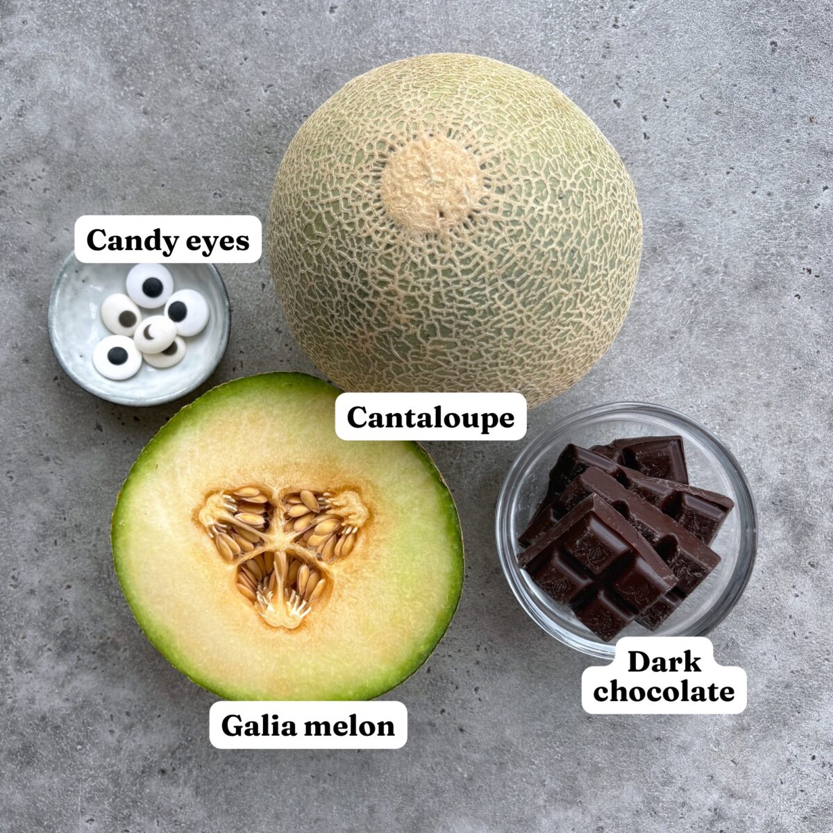 A whole cantaloupe, a halved Galia melon, a bowl of candy eyes and dark chocolate squares.