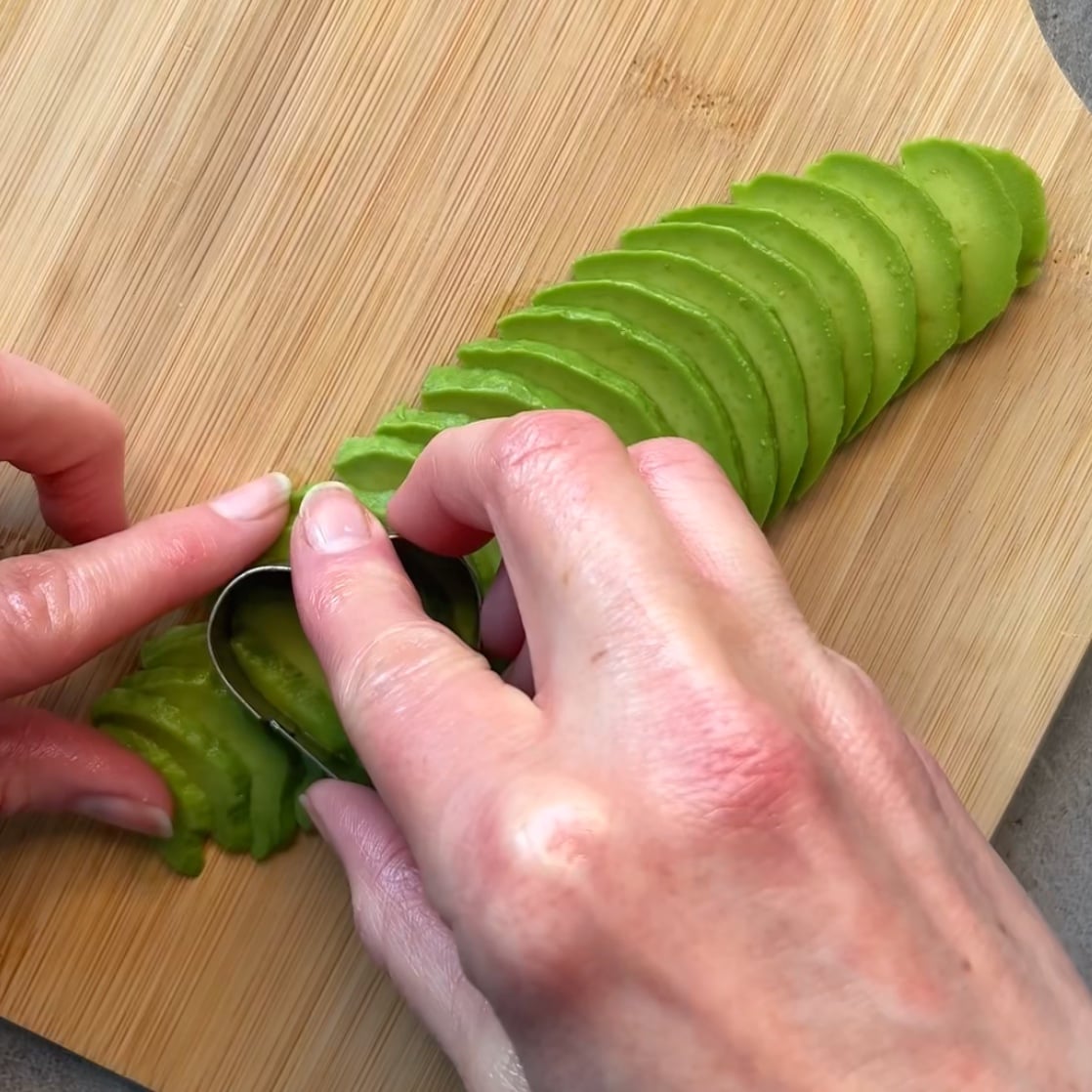 Hands arranging thin slices of avocado on a wooden cutting board and using a small cookie cutter. 