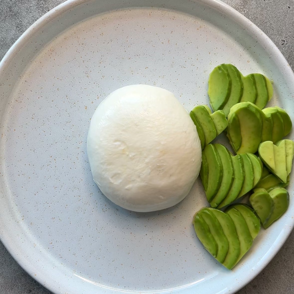 A ball of burrata cheese sits next to sliced avocado pieces cut into heart shapes, on a round white plate.