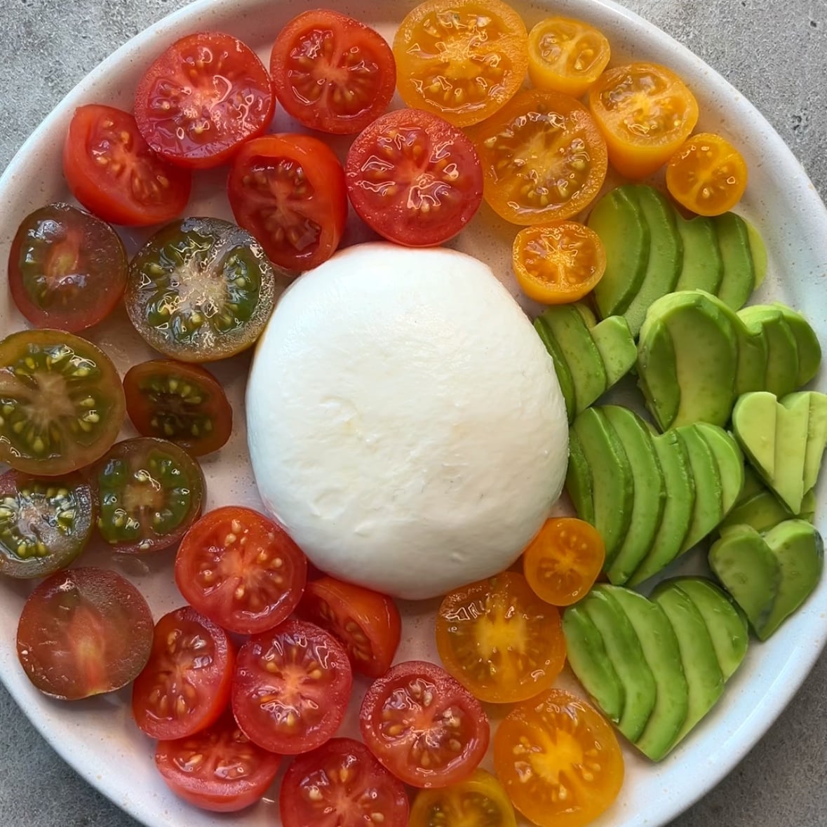 A plate with sliced red, yellow, and green tomatoes, sliced avocado, and a round ball of burrata cheese in the center.