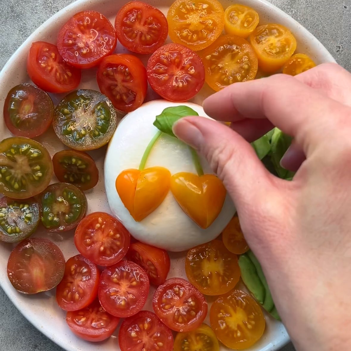 A hand places basil leaves next to two orange tomato halves shaped like hearts on creamy burrata, surrounded by colorful halved cherry tomatoes.