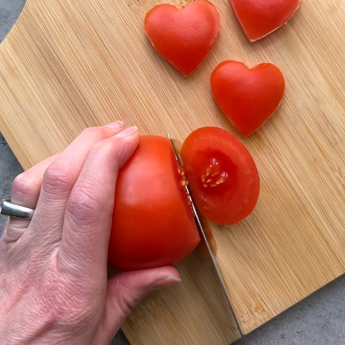 A hand slices a tomato on a wooden cutting board. Three heart-shaped tomato pieces are placed nearby.