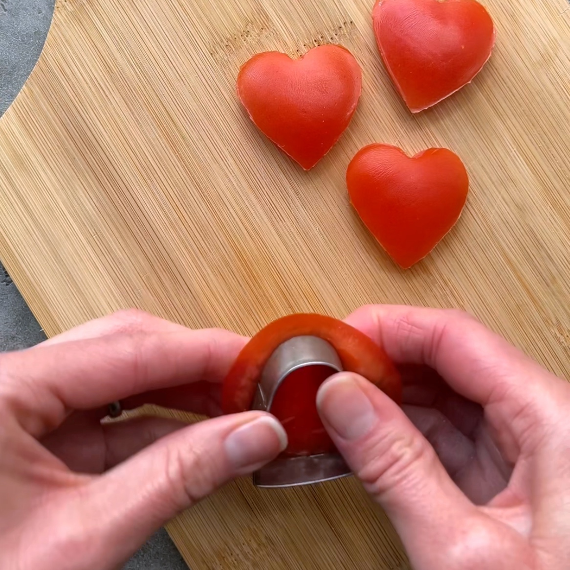 Hands using a heart-shaped cutter to cut a slice of tomato.
