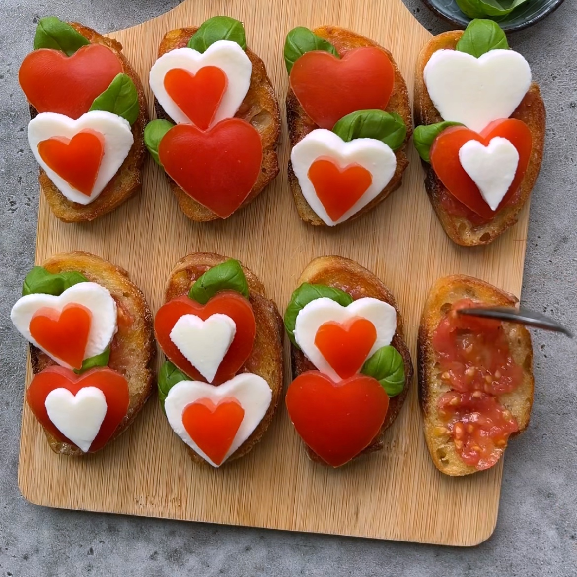 Slices of toasted bread topped with basil, mozzarella, and heart-shaped tomato pieces are arranged on a wooden board. A hand spoons a tomato mixture onto a crostini. 