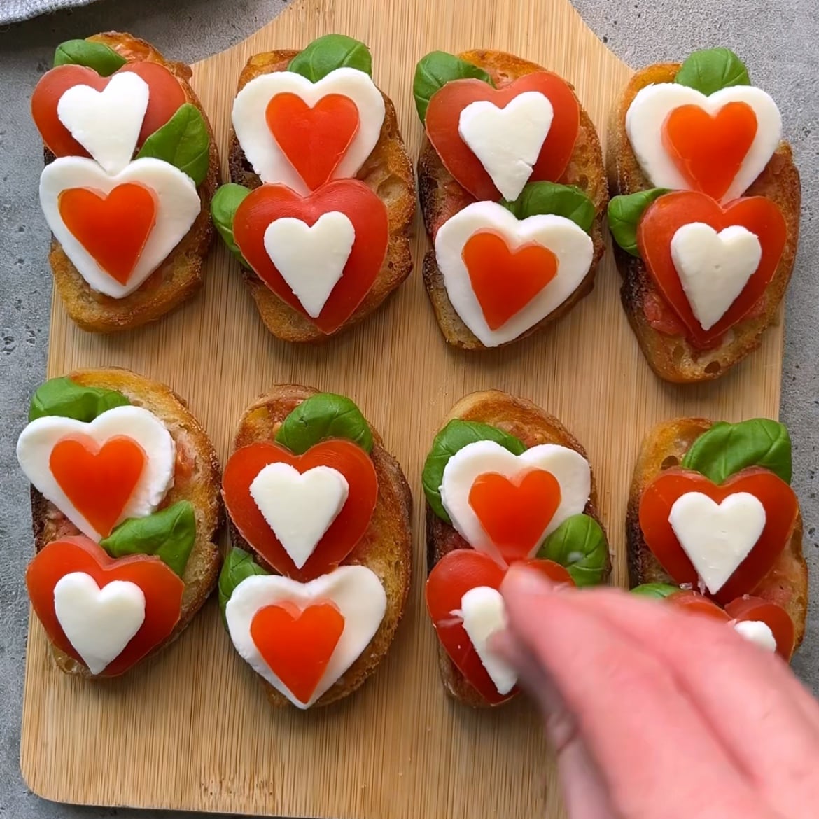Slices of bread topped with basil, and heart-shaped tomato and mozzarella are arranged on a wooden board. A hand sprinkles sea salt on one piece.