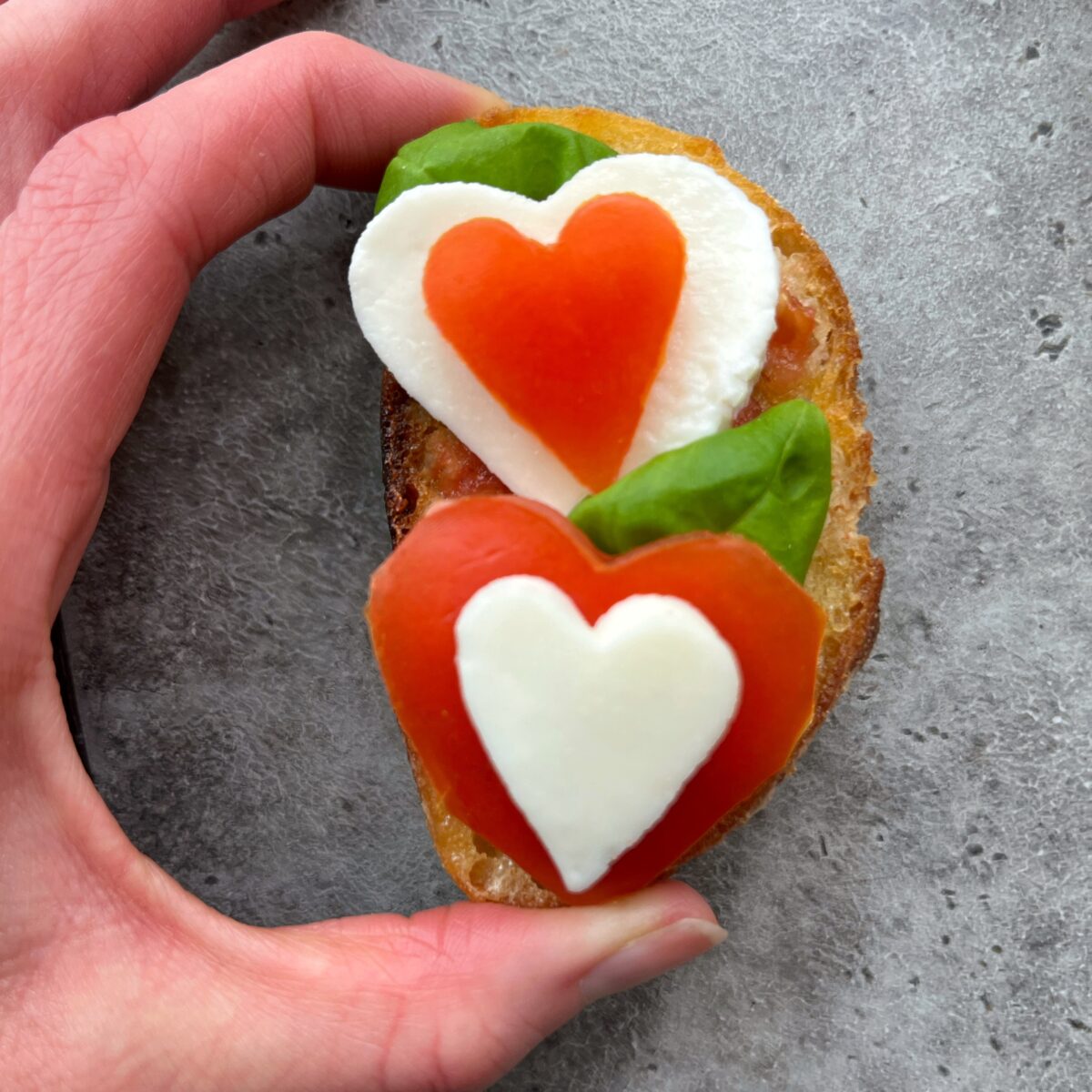 A hand holds a piece of bread topped with heart-shaped slices of tomato and mozzarella, fresh basil leaves, on a gray surface.