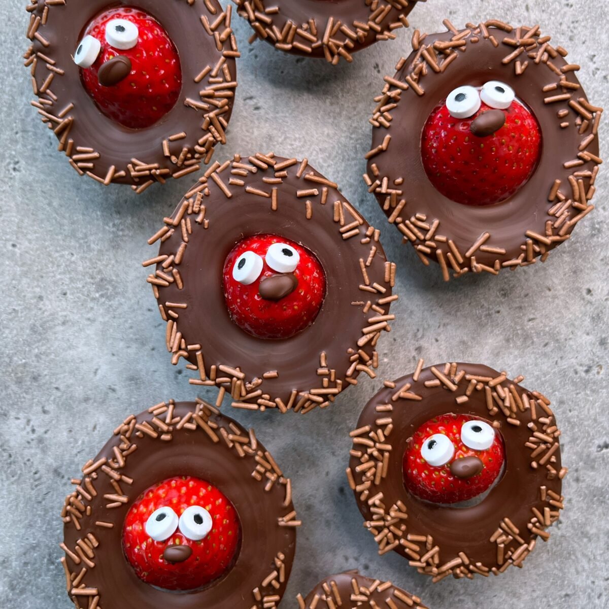 Chocolate-covered strawberry bites topped with candy eyes, brown sprinkles, and cartoon-like facial expressions arranged on a gray surface.