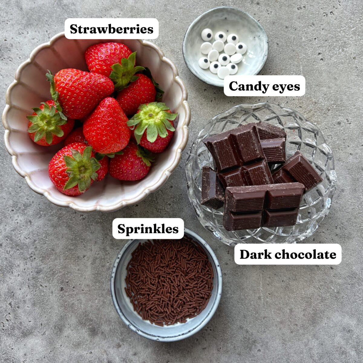 Ingredients for Valentine Chocolate Hedgehogs including a white scalloped bowl of strawberries, a glass bowl of dark chocolate pieces, candy eyes, and sprinkles. All labelled. 