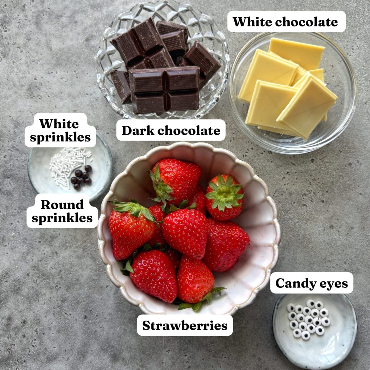 A bowl of strawberries, dark chocolate, white chocolate, candy eyes, white sprinkles, and round sprinkles arranged on a countertop.
