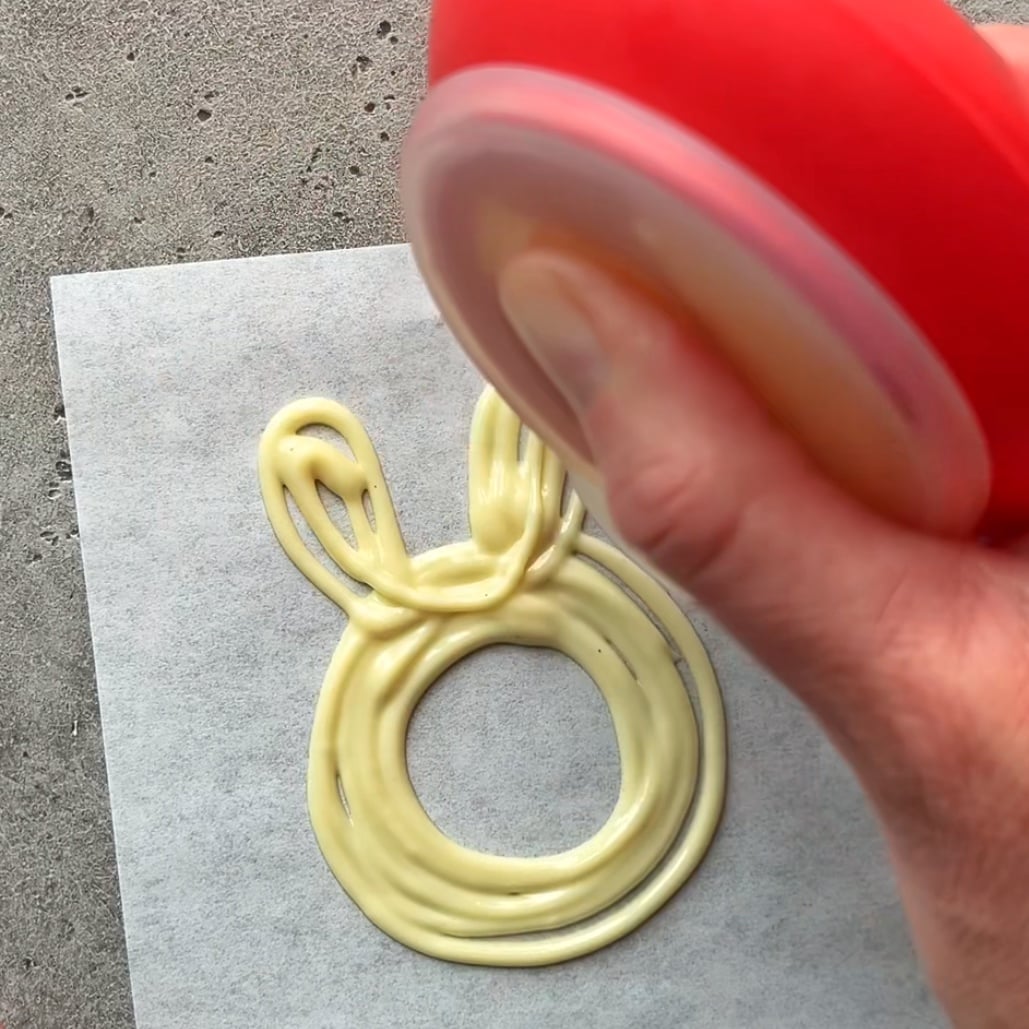 A hand squeezes white chocolate from a red dispenser onto parchment paper, forming a bunny shape with a circular face and two ears.