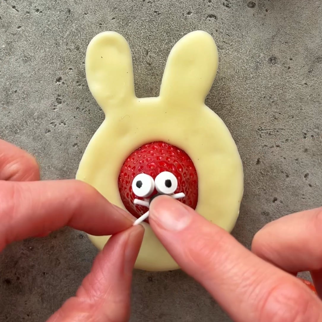 A hand outlines a Chocolate Easter Bunny shape with chocolate on parchment paper next to two strawberry-filled white chocolate bunny treats with candy eyes and whiskers.