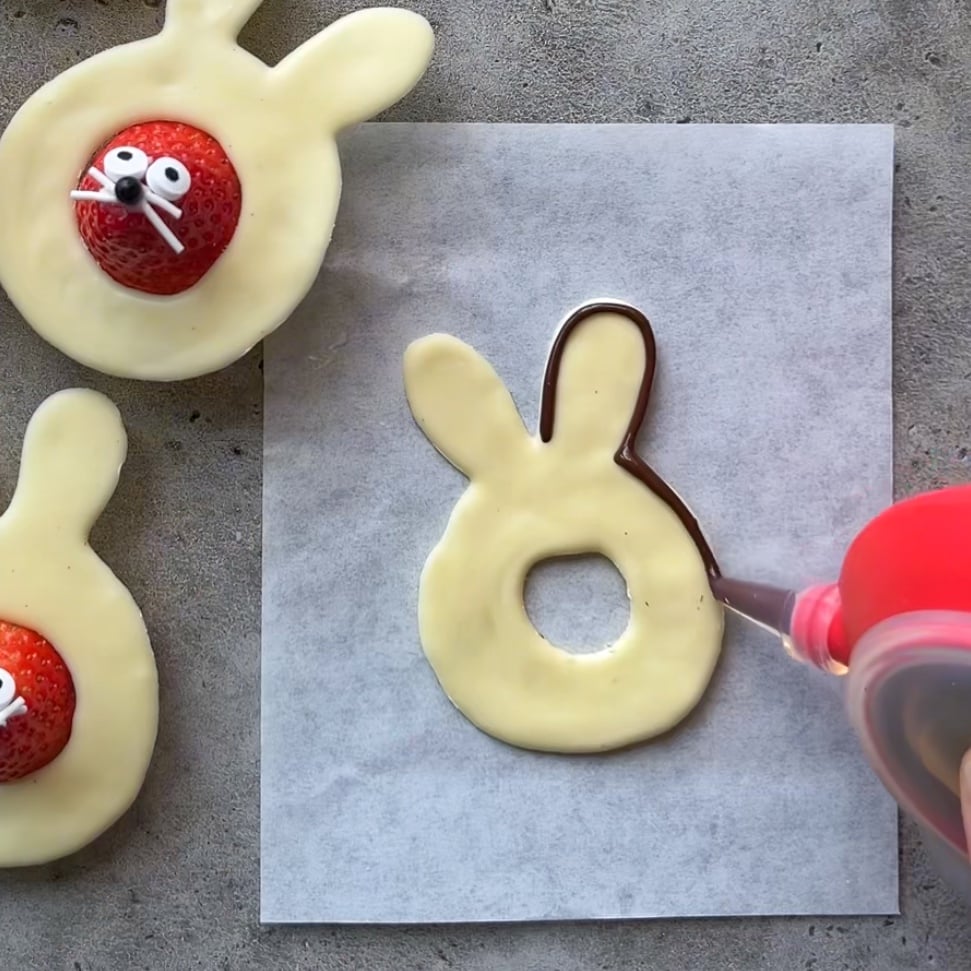 Hands decorate a strawberry with candy eyes and whiskers, placing it in the center of a white chocolate Easter Bunny.