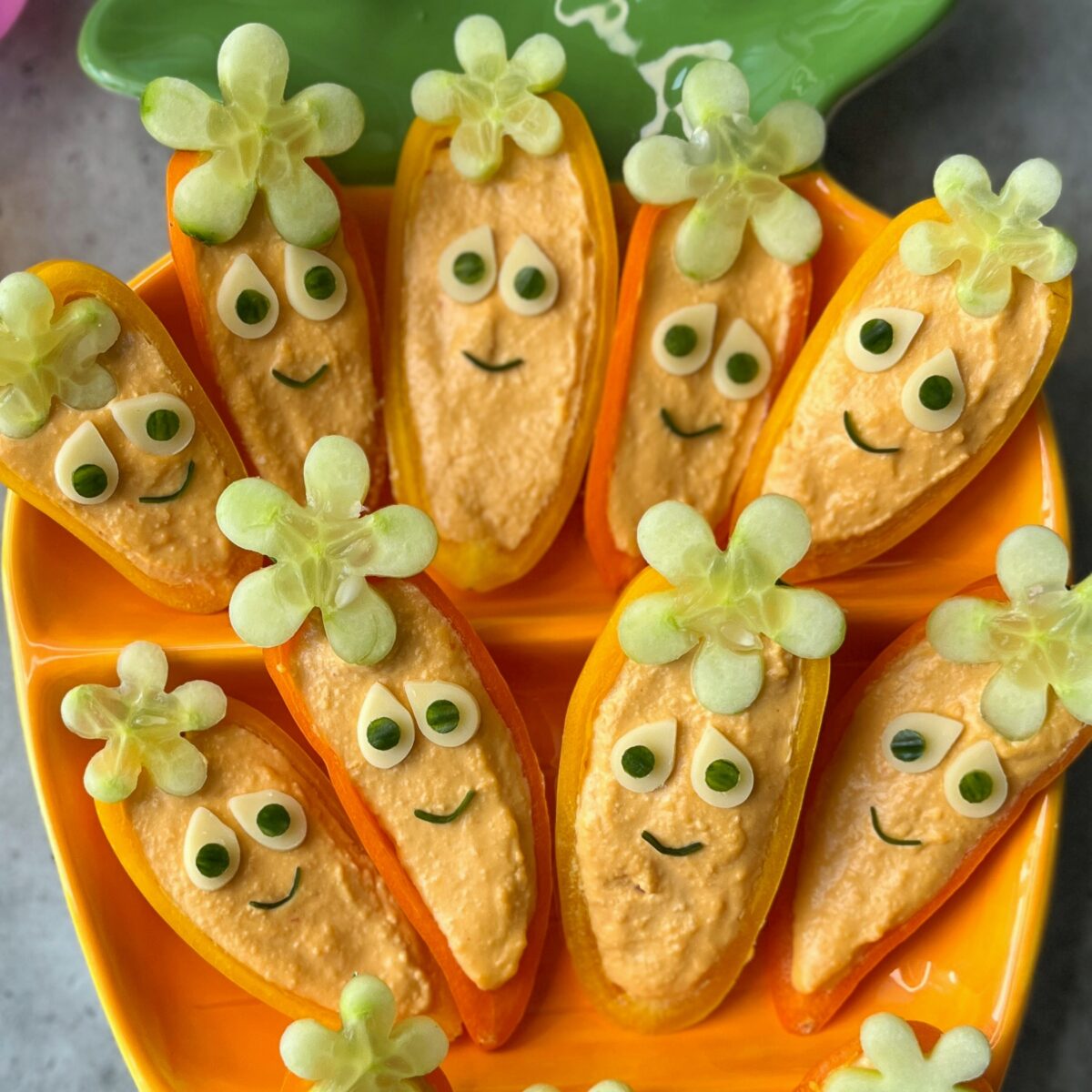 Orange bell pepper halves filled with hummus and topped with cucumber made to look like carrots, arranged on a bright yellow plate.