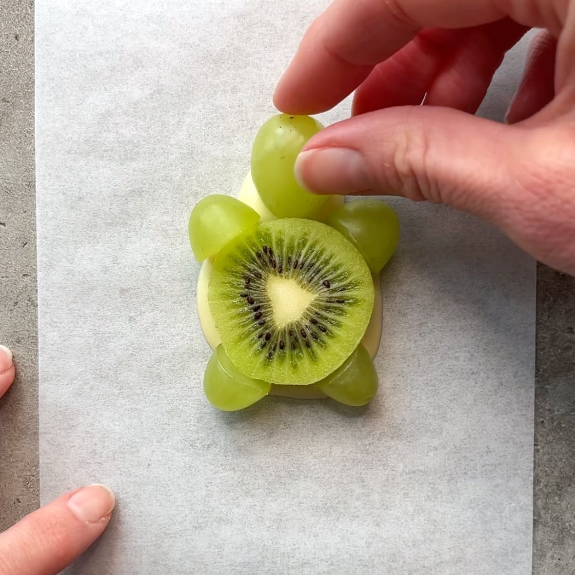 A hand places a green grape 'head' onto white chocolate.