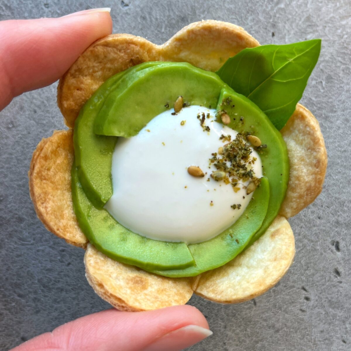 A hand holds a flower-shaped bite made of baked tortilla, sliced avocado, and a dollop of whipped feta.