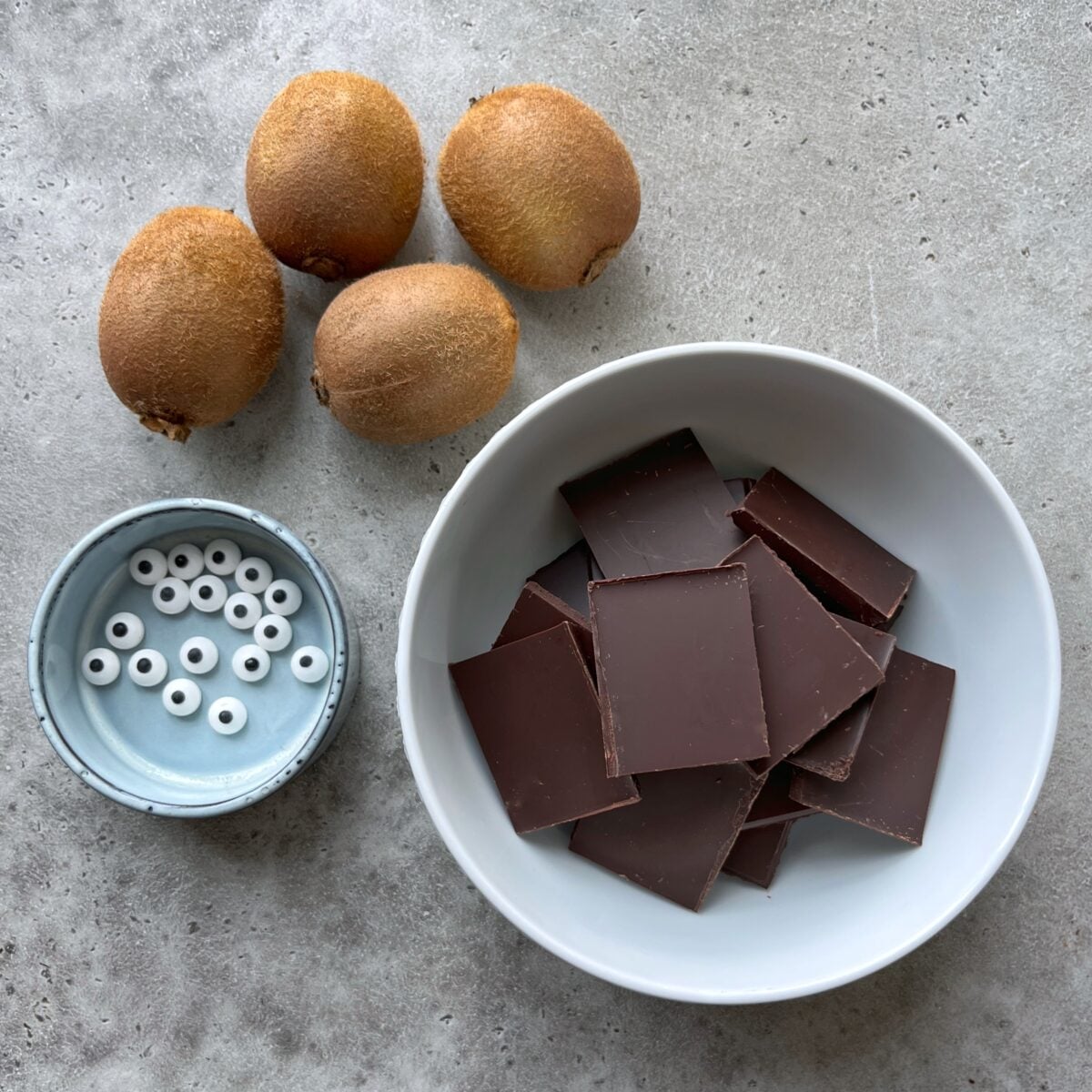 Four whole kiwis, a bowl of chocolate squares, and a small dish of candy eyes on a gray surface.