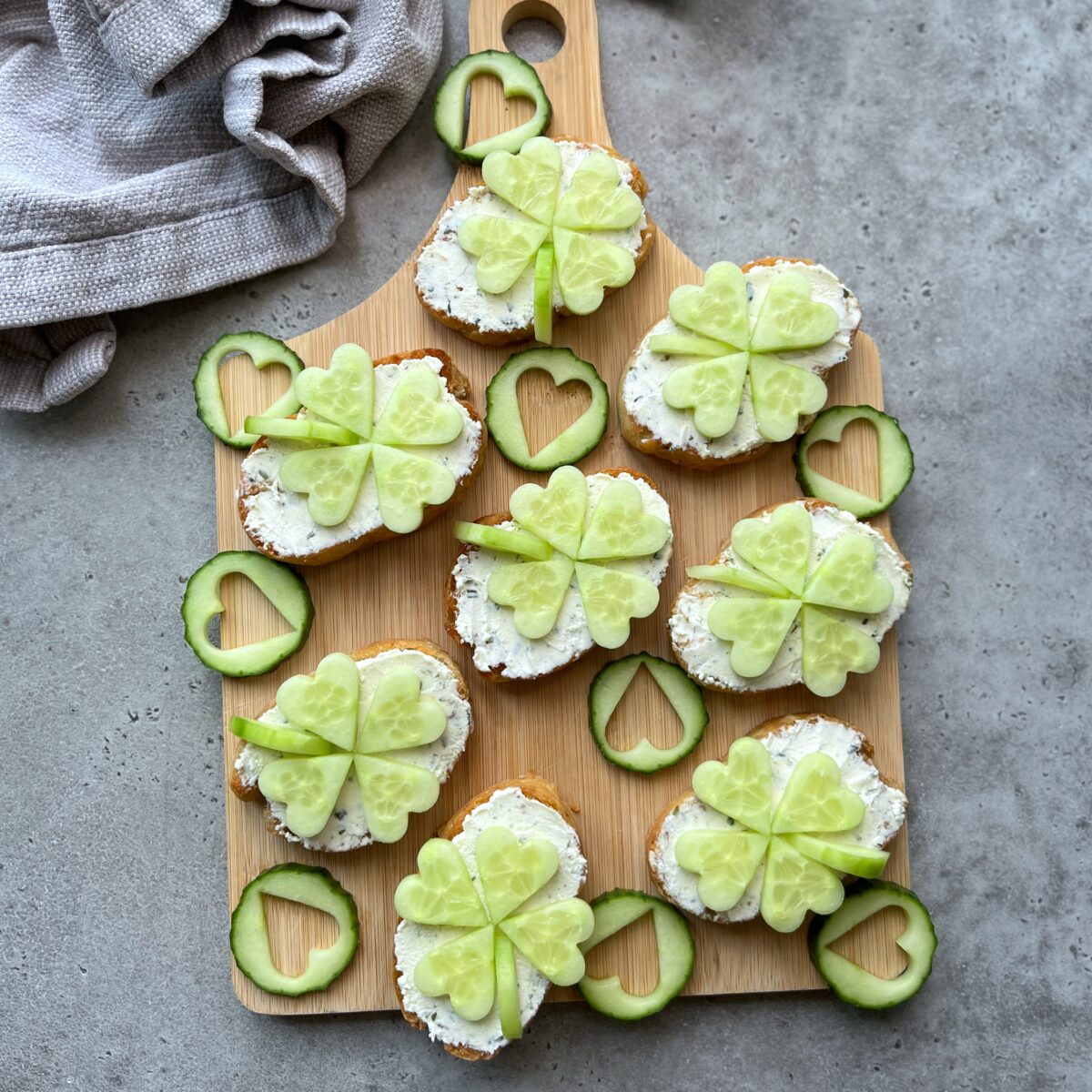 Small crostini with cream cheese topped with cucumber slices shaped like four-leaf clovers, arranged on a wooden serving board.