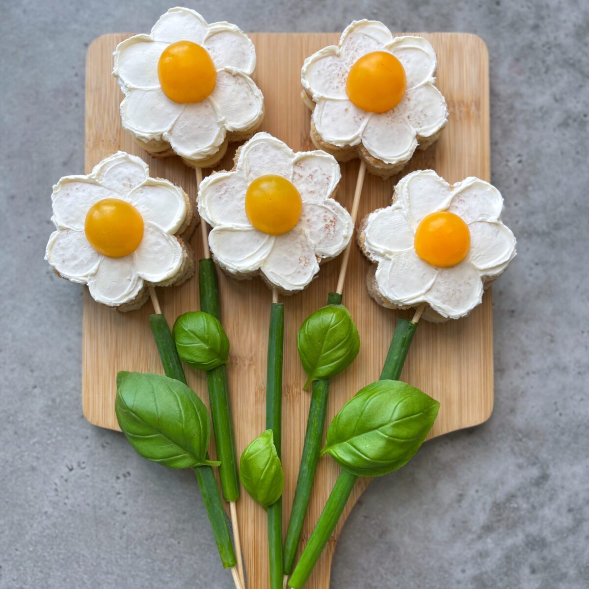 Five sandwiches decorated as flowers with cream cheese petals and yellow cherry tomato centres are arranged on a wooden board, with fresh basil resembling stems and leaves.