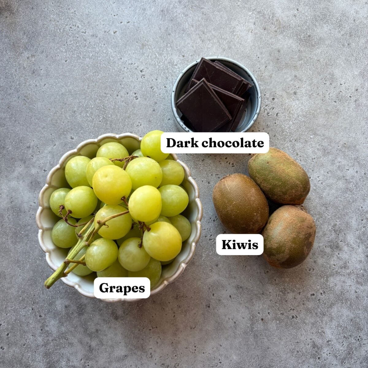 A bowl of green grapes, three kiwis, and dark chocolate squares.