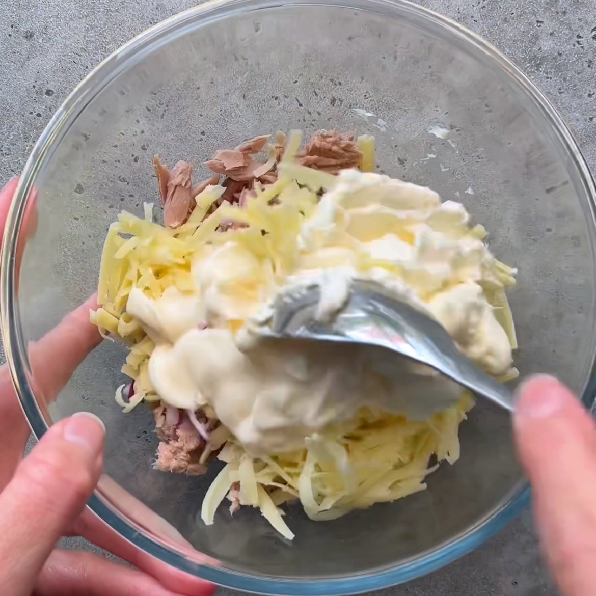 A person mixes shredded red onion, cheddar cheese, tuna, cream cheese, and mayonnaise in a glass bowl with a spoon.